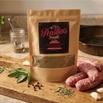 Parillo's Breakfast Sausage seasoning pack on a wooden cutting board beside fresh raw sausage links, sage leaves, thyme, rosemary, garlic cloves, black peppercorns, and a spice jar in a warm rustic kitchen