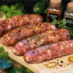Fresh raw Parillo's Retail Breakfast Bulk sausage links seasoned with crushed chili flakes and dried herbs on a wooden cutting board, surrounded by fresh basil, oregano, thyme, roasted garlic, dried red chili peppers, and jars of crushed chili and black pepper on a dark background