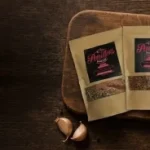 Three Parillo Sausage seasoning packets displayed on a wooden cutting board surrounded by garlic, fresh herbs, spices, and grilled chicken wings on a dark rustic background.