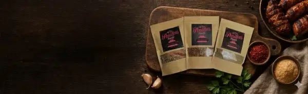 Three Parillo Sausage seasoning packets displayed on a wooden cutting board surrounded by garlic, fresh herbs, spices, and grilled chicken wings on a dark rustic background.