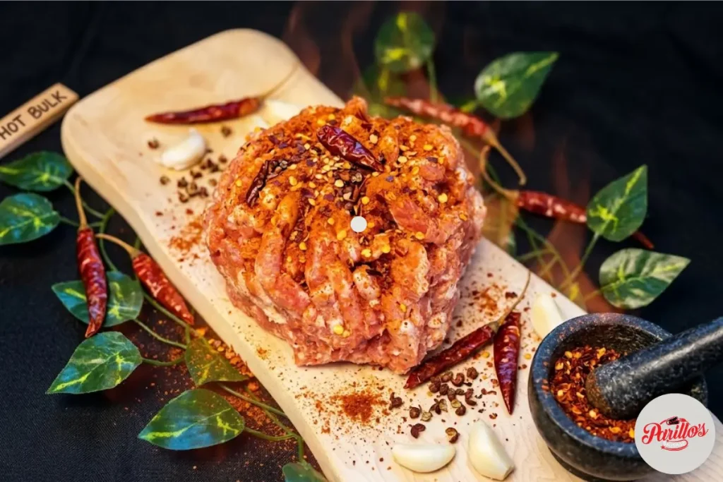 Parillo's Hot Bulk ground sausage meat heavily seasoned with dried red chili peppers and chili flakes on a wooden cutting board with a Hot Bulk label, surrounded by garlic cloves, green leaves, chili powder and a stone mortar filled with crushed spices on a dark background