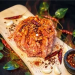 Parillo's Hot Bulk ground sausage meat heavily seasoned with dried red chili peppers and chili flakes on a wooden cutting board with a Hot Bulk label, surrounded by garlic cloves, green leaves, chili powder and a stone mortar filled with crushed spices on a dark background