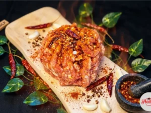 Parillo's Hot Bulk ground sausage meat heavily seasoned with dried red chili peppers and chili flakes on a wooden cutting board with a Hot Bulk label, surrounded by garlic cloves, green leaves, chili powder and a stone mortar filled with crushed spices on a dark background
