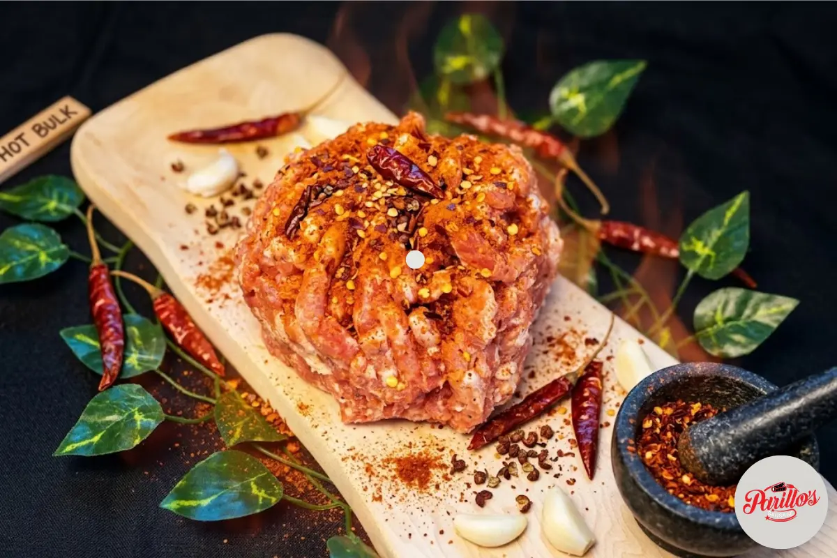 Parillo's Hot Bulk ground sausage meat heavily seasoned with dried red chili peppers and chili flakes on a wooden cutting board with a Hot Bulk label, surrounded by garlic cloves, green leaves, chili powder and a stone mortar filled with crushed spices on a dark background