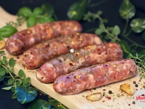 Fresh raw Parillo's hot link sausages seasoned with red chili flakes and mixed herbs, laid on a wooden cutting board surrounded by fresh oregano, thyme, basil, roasted garlic cloves and mixed peppercorns on a dark background