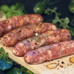Five raw breakfast link sausages from Pirillo's, seasoned with red pepper flakes and herbs, arranged on a wooden cutting board surrounded by fresh basil, thyme, oregano, whole peppercorns, and roasted garlic cloves against a dark background.