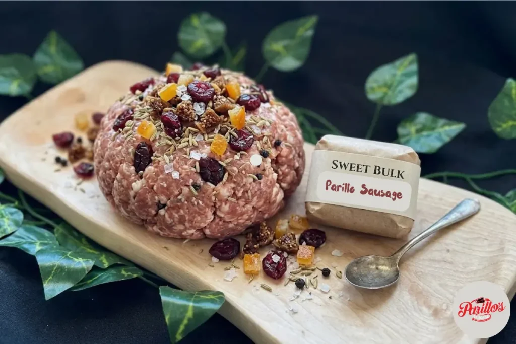 Parillo's Sweet Bulk ground sausage meat topped with dried cranberries, diced apricots, fennel seeds and coarse salt on a wooden cutting board with a Sweet Bulk Parillo Sausage label and vintage spoon, surrounded by green ivy leaves on a dark background