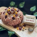 Parillo's Sweet Bulk ground sausage meat topped with dried cranberries, diced apricots, fennel seeds and coarse salt on a wooden cutting board with a Sweet Bulk Parillo Sausage label and vintage spoon, surrounded by green ivy leaves on a dark background