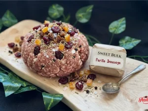 Parillo's Sweet Bulk ground sausage meat topped with dried cranberries, diced apricots, fennel seeds and coarse salt on a wooden cutting board with a Sweet Bulk Parillo Sausage label and vintage spoon, surrounded by green ivy leaves on a dark background