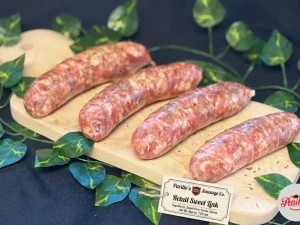 Four fresh Parillo's Retail Sweet Link sausages made with sweet pork, fennel and spices, displayed on a wooden cutting board with a product label tag, surrounded by green vine leaves on a dark background